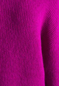 Fuchsia stickad tröja med ribbat mönster. Materialet verkar tjockt och varmt, med en kontinuerlig yta och subtila variationer.