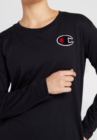 T-shirt a maniche lunghe nera in cotone, con logo circolare ricamato con accento rosso. Tessuto liscio, collo a giro, maniche aderenti.