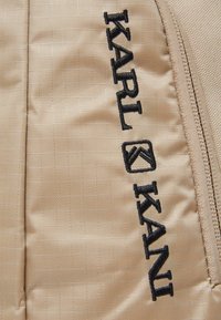 Beżowa, pikowana tkanina z teksturowanym wzorem w kratkę i czarnym haftowanym logo "KARL KANI" wzdłuż szwu bocznego. Widoczny detal zamka.