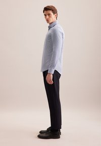 Camicia a maniche lunghe azzurro chiaro con un piccolo motivo a quadretti, abbinata a pantaloni scuri eleganti e scarpe nere, posata di lato.