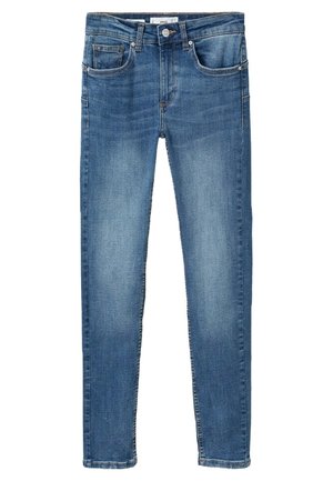 Persoon draagt een beige trenchcoat, wit t-shirt, blauwe skinny jeans en zwarte ballerina's, staat met handen in de zakken.