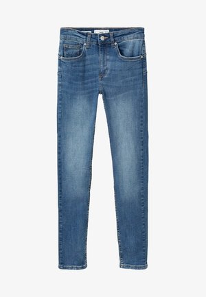 Personne portant un trench beige, un t-shirt blanc, un jean skinny bleu et des ballerines noires, debout avec les mains dans les poches.