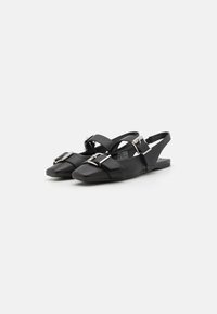 Zapatos de piel negra tipo slingback con suela plana, que cuentan con una prominente hebilla plateada y correas cruzadas para un ajuste seguro.