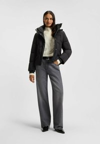 Svart puffjacka med hög krage, i kombination med en ljus, texturerad tröja och vida grå jeans. Svarta skor fulländar outfiten.