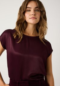 Burgundy silk blouse with a relaxed fit, short cap sleeves, round neckline, and subtle sheen, paired with dark fabric textured pants.