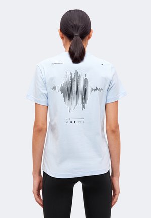 Femme aux cheveux attachés porte un t-shirt bleu clair avec des icônes noires d’onde sonore et de lecteur de musique dans le dos.