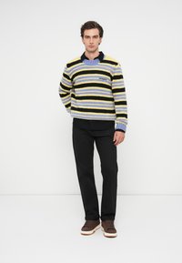 Pull rayé en jaune, noir et violet ; texture tricotée. Porté par-dessus une chemise noire à col, associé à un pantalon noir et des baskets marron.