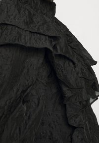 Tessuto nero con texture, dettagliato da strati di rouches e motivi intricati, dotato di un colletto alto. La superficie appare leggermente lucida e strutturata.