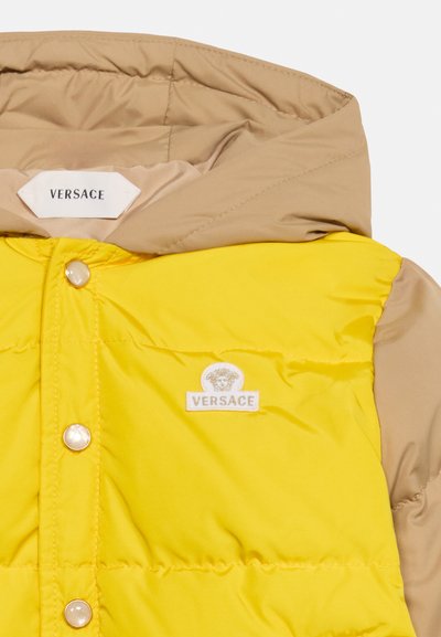Section de doudoune Versace jaune et beige avec boutons-pression, écusson logo Méduse et étiquette Versace à l'intérieur de la capuche beige.