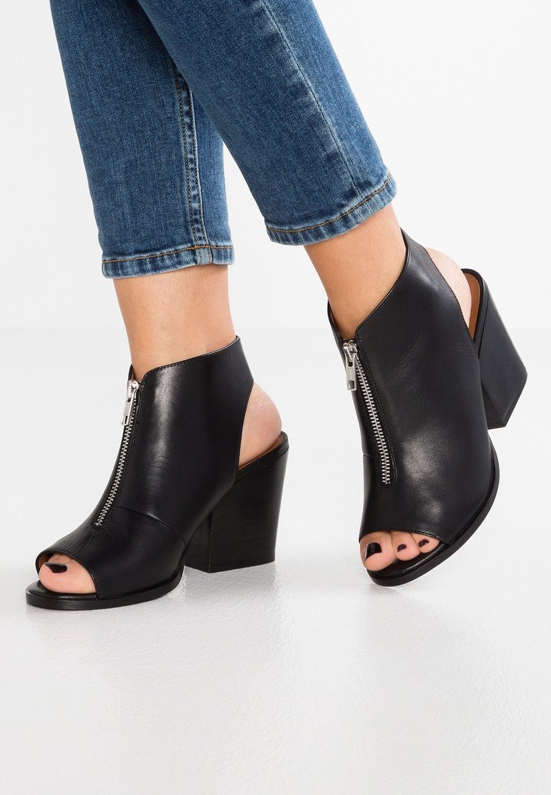 Czarne skórzane ankle boots z odkrytymi palcami oraz zamkiem z przodu, noszone z niebieskimi podwiniętymi dżinsami na białej podłodze.