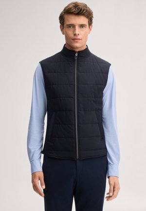Uomo che indossa un gilet trapuntato nero con zip sopra una camicia azzurra a maniche lunghe, in piedi davanti a uno sfondo chiaro e uniforme.
