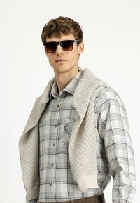 Mężczyzna w jasnobeżowym dzierganym swetrze zarzuconym na ramiona, w koszuli w kratę w stonowanych kolorach oraz w ciemnych okularach przeciwsłonecznych.