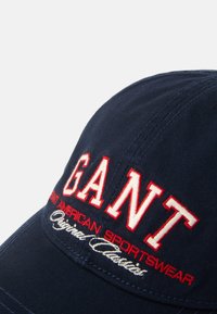 Tummansininen baseball-lippis, joka on valmistettu puuvillasta ja jossa on punaisella ja valkoisella brodeerattu teksti "GANT" ja "Original Classics" edessä.