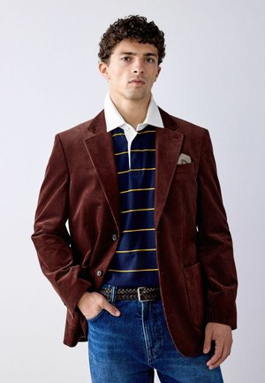 Young man wearing a brown corduroy blazer over a navy blue striped polo shirt with a white collar and blue jeans, standing with one hand in pocket.