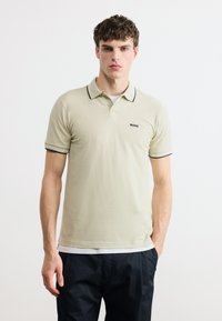 Jeune homme portant un polo beige avec des bordures noires et le logo "BOSS", associé à un pantalon noir, debout devant un fond blanc uni.