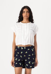 Hvit cropped bluse med blonder og knappelukking, kombinert med marineblå shorts med sitronmønster. Teksturert stoff og volanger.