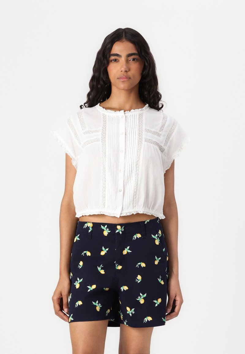 Hvit cropped bluse med blonder og knappelukking, kombinert med marineblå shorts med sitronmønster. Teksturert stoff og volanger.