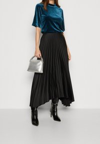 Tyrkysový sametový top s krátkými rukávy, v kombinaci s černou plisovanou sukní a černými vyvýšenými botami. Drží stříbrnou kabelku s řetízkovým úchytem.