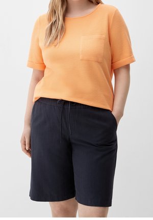 Kvinde iført ferskenfarvet kortærmet tekstureret top med brystlomme og sorte knælange shorts med snøre, hænder afslappet ned langs siderne og i lommen.