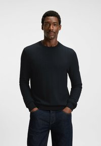 Black textured knit sweater with a herringbone pattern, featuring a crew neck and ribbed cuffs, paired with dark denim jeans.