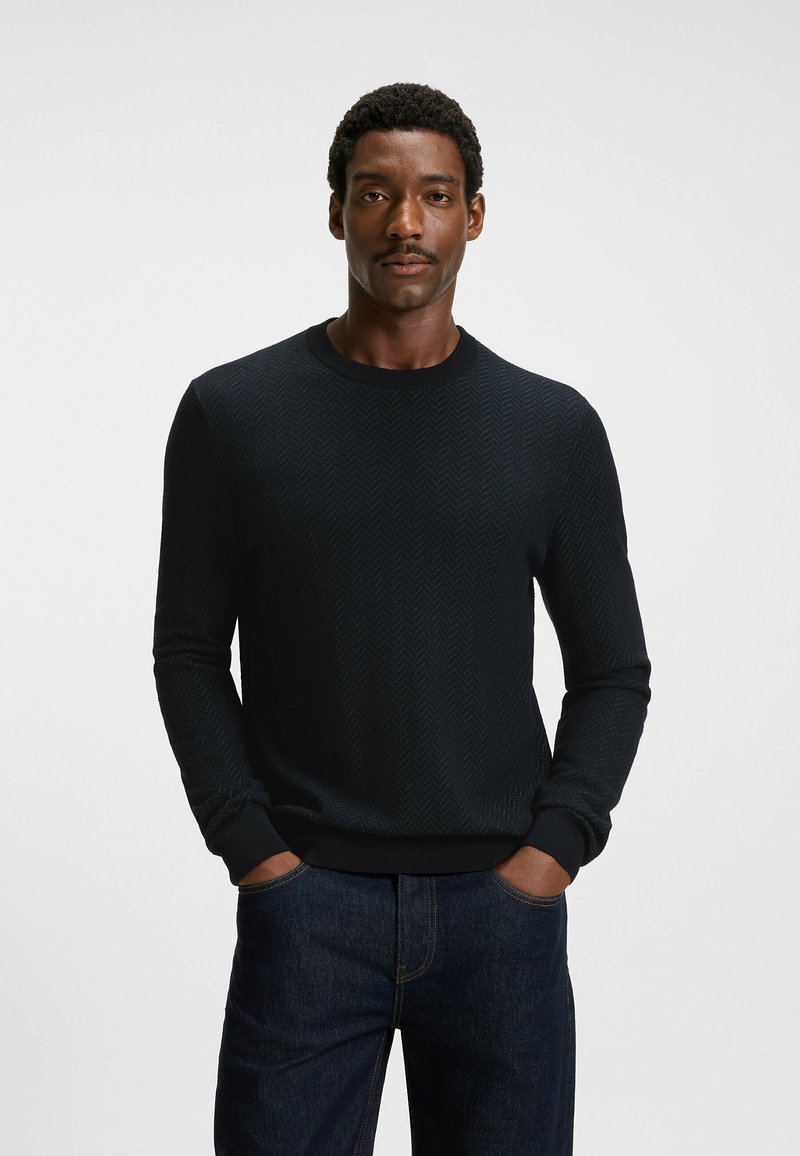 Black textured knit sweater with a herringbone pattern, featuring a crew neck and ribbed cuffs, paired with dark denim jeans.