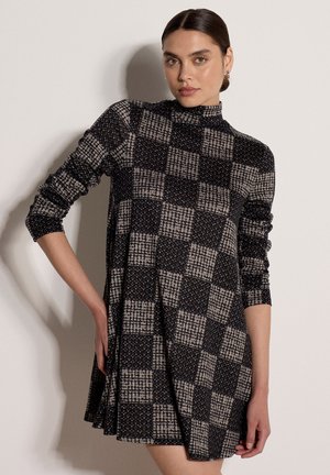 Femme aux cheveux foncés portant une robe à manches longues à carreaux noirs et gris, debout avec une main sur la hanche, sur un fond uni.