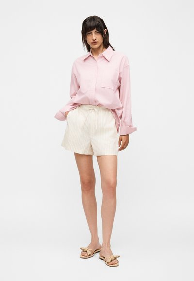 Γυναίκα στέκεται με τα χέρια στις τσέπες φορώντας μια φαρδιά ροζ ριγέ πουκάμισο, κρεμ σορτς και μπεζ σανδάλια με διακοσμητικά φιόγκους.