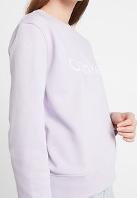 Sudadera color lila claro con cuello redondo, mangas largas y el logo "Calvin Klein" en blanco en el pecho. Textura de tela suave.