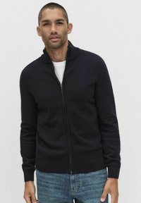 Granatowy kardigan zapinany na zamek z wysokim kołnierzem, �ściągaczami przy mankietach i u dołu; wykonany z miękkiego materiału dzianinowego, w połączeniu z jasnoniebieskimi dżinsami.