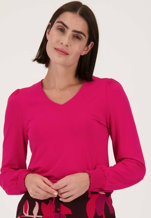 Haut à manches longues fuchsia avec un décolleté en V, détails froncés sur les manches et texture lisse. Porté avec une jupe à motif floral.