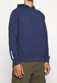Navy blue pullover hoodie made of soft fabric, featuring a kangaroo pocket and red logo accent. Includes ribbed cuffs and hem.