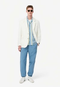 Blazer in lino bianco sopra una camicia azzurra a fantasia, abbinata a pantaloni stile jogger azzurri e sneaker bianche con dettagli verdi.