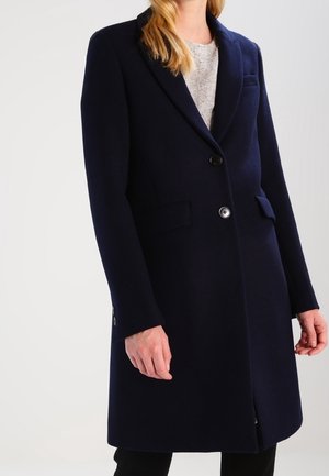 Frau trägt einen langen, dunkel marineblauen, maßgeschneiderten Mantel mit zwei Knöpfen und Klappentaschen über einem hellgrauen Oberteil.