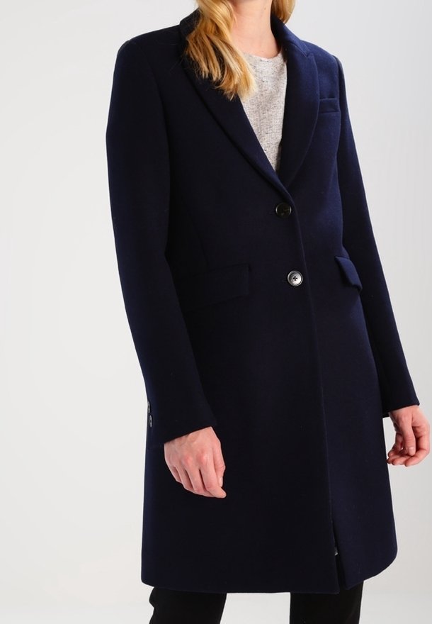 Femme portant un long manteau ajusté bleu marine foncé avec deux boutons et des poches à rabat, par-dessus un haut gris clair.