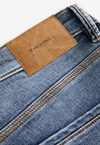 Svetlomodrá rifľovina s oranžovým prešívaním a obdĺžnikovou hnedou koženou nášivkou s nápisom "DENIM SUPPLY Co." na páse.