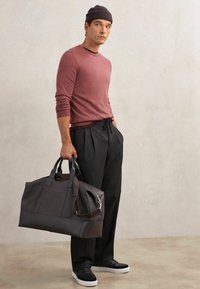 Ciemna skórzana torba podróżna z dwoma górnymi uchwytami i regulowanym paskiem, z teksturowanymi panelami i zamkiem błyskawicznym w kolorze srebrnym. Ubranie i czapka w ciemnym kolorze.