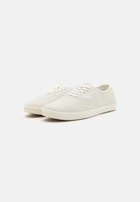 Light beige suede sneakers with a low-top design, round toe, and white laces. Features a textured surface and a minimalist aesthetic.