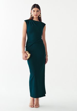 Femme portant une robe longue sans manches vert foncé, tenant une pochette dorée, avec des boucles d'oreilles et un bracelet dorés, debout devant un fond blanc.