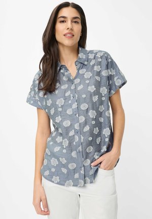 Femme aux longs cheveux bruns portant une chemise bleue à manches courtes avec des motifs de fleurs blanches et un pantalon blanc, main dans la poche.