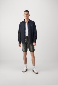 Un tânăr stă îmbrăcat cu o cămașă neagră cu butoni peste un maiou alb, pantaloni scurți din denim închis, răsuciți, șosete albe și adidași albi.