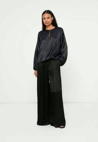 Svart satengbluse med langermet, rund hals og glidelåsdetalj foran, kombinert med vide svarte bukser som viser en jevn tekstur.