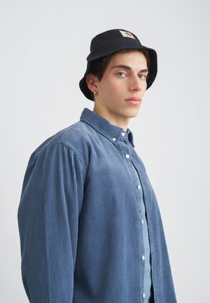 Kék kordbársony ing gombos záródással, fekete kerek kalappal párosítva, amely logóval díszített. Sima textúra és laza fit.