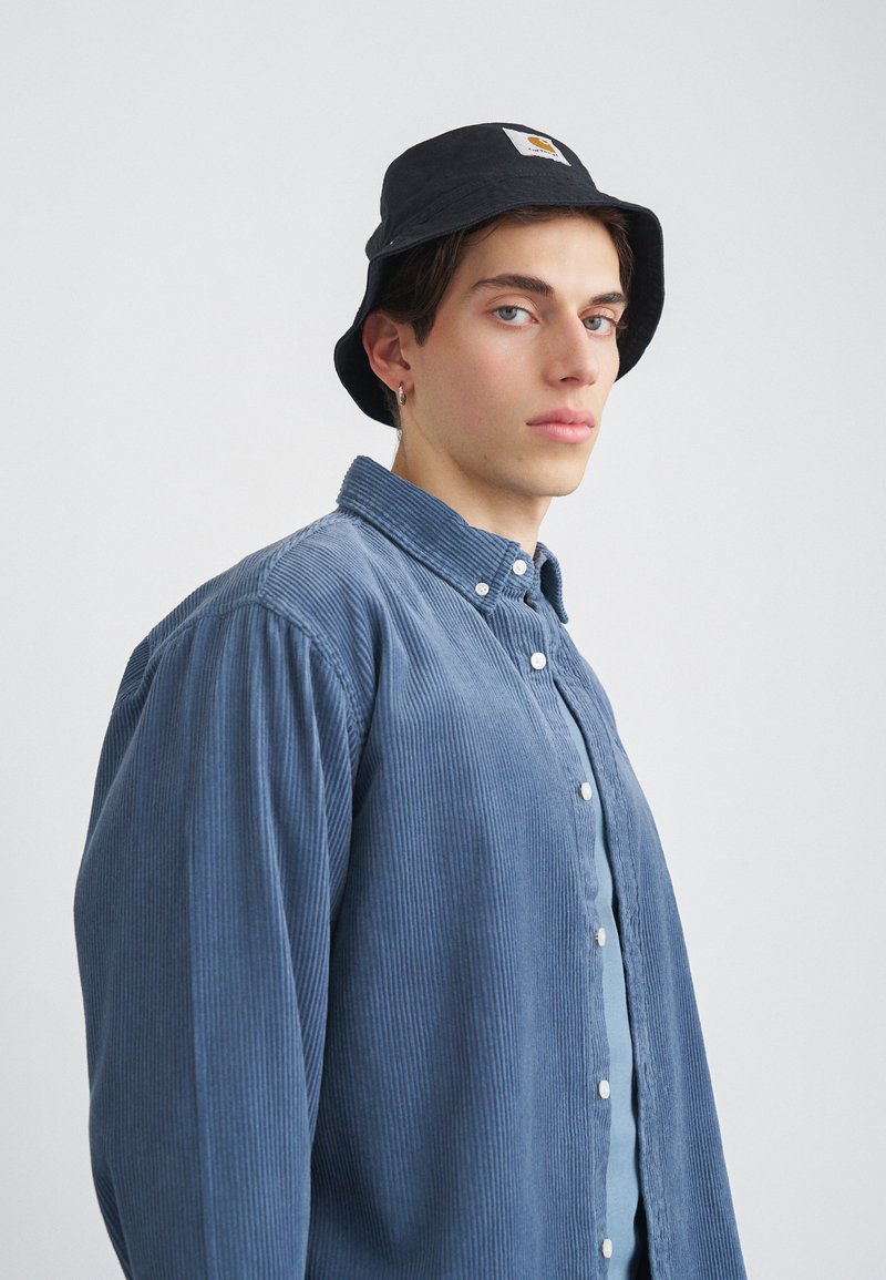 Modrá korduroy košeľa s gombíkovým zap�ínaním, skombinovaná s čiernym vedierkovým klobúkom s nášivkou. Hladká textúra a uvoľnený strih.