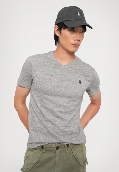 Γκρι κοντομάνικο T-shirt με V-λαιμόκοψη και λεπτή υφή, διακοσμημένο με ένα μικρό σκούρο λογότυπο στο στήθος, συνδυασμένο με σκούρο καπέλο και πράσινα cargo σορτς.