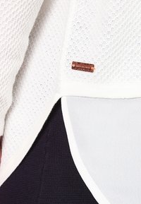 Top de tela blanca de malla con un diseño texturizado y etiqueta en tono cobre. Presenta un estilo de capas con una tela oscura contrastante debajo.
