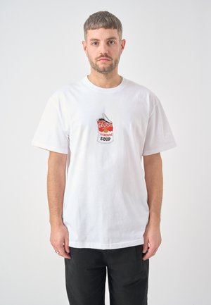 Mann mit kurzen Haaren und Bart, der ein weißes T-Shirt mit einer rot-weißen Suppendosen-Grafik und schwarze Hose vor einem einfarbigen Hintergrund trägt.