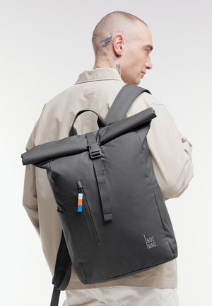 Homme avec des tatouages portant une veste beige, portant un sac à dos noir à fermeture roulante avec une fermeture éclair frontale et le logo "GOT BAG".