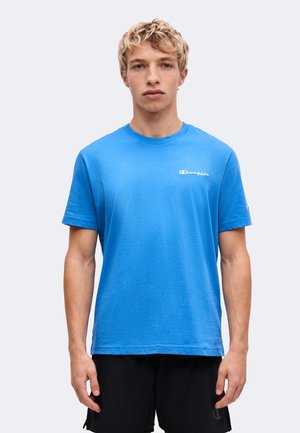 Jeune homme aux cheveux blonds courts portant un t-shirt bleu Champion et un short noir, debout devant un fond clair et uni.