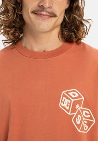 Hombre con cabello rizado y bigote, vistiendo una camisa de color óxido con dos dados contorneados en blanco con letras y símbolos en el pecho.