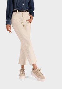 Donna che indossa pantaloni cropped beige con orli sfrangiati, sneakers beige con zeppa, una cintura nera e una camicia a maniche lunghe blu scuro.
